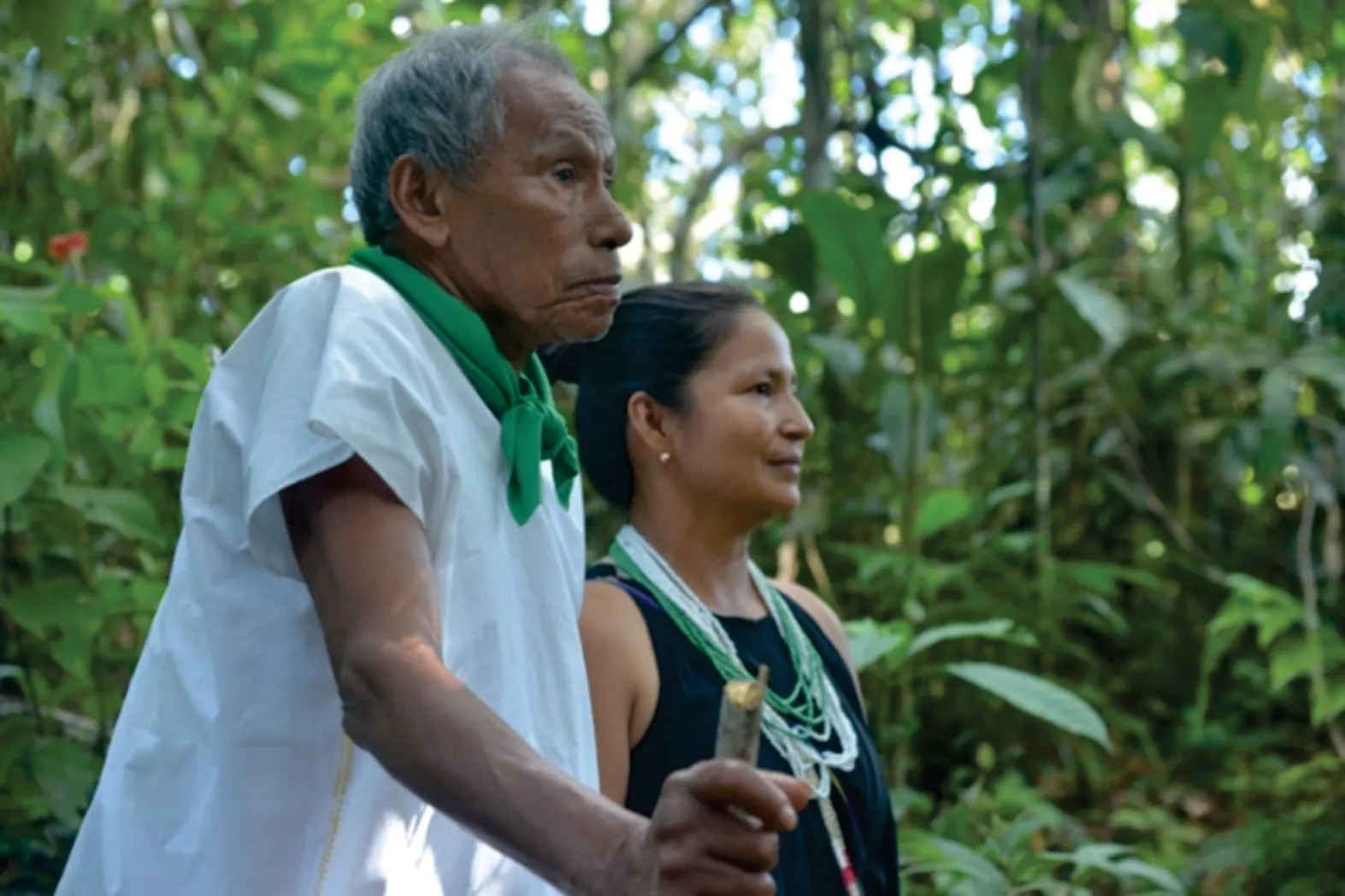 Mayor con camisa blanca y pañuelo verde junto a una mujer con collares, en la selva.