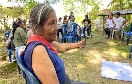 Mayor sentada señalando con la mano durante una reunión al aire libre con sillas plásticas y papelógrafos.
