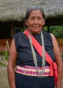 Retrato de una mayor con blusa negra, collares, chumbe y banda roja frente al tambo (línea de tiempo).