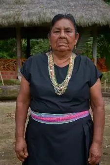 Retrato de una mayor con blusa negra, collares y chumbe rosado frente al tambo (línea de tiempo).