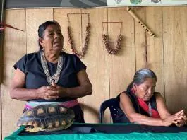 Dos mayoras en un salón de madera con collares colgados en la pared; una con caparazón de tortuga (línea de tiempo).