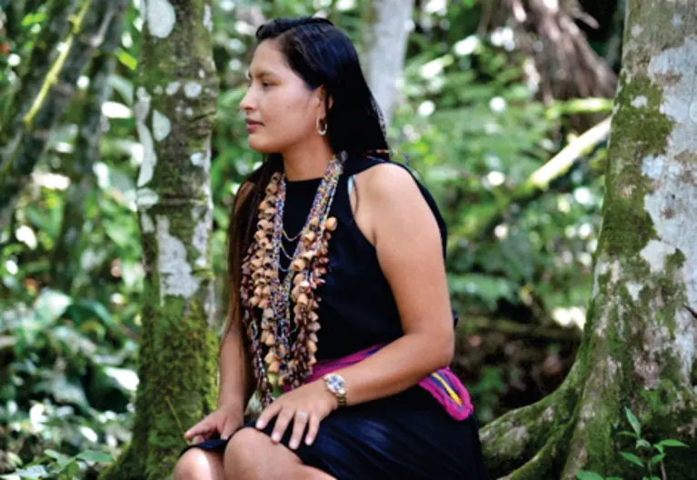 Joven mujer con vestido tradicional, collares de semillas y chumbe, sentada junto a un árbol (vestido tradicional).