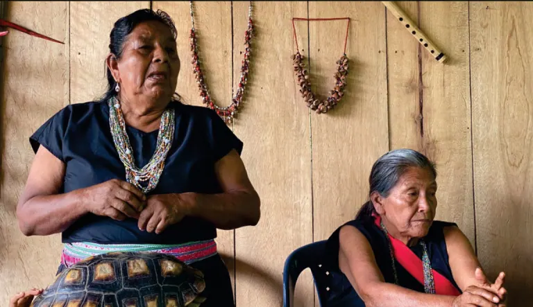 Dos mayoras en un salón de madera con collares colgados en la pared; una sostiene un caparazón de tortuga (vestido tradicional).
