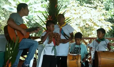 Hombre tocando guitarra y niños tocando bombos con collares de semillas (música e instrumentos).