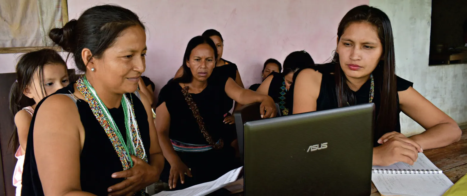 Mujeres trabajando con un computador portátil y cuadernos en una mesa; una niña al lado (justicia propia).
