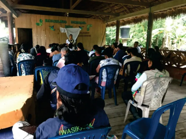 Asamblea en un salón de madera con muchas personas sentadas de espaldas; una con chaleco Wasikama (educación).
