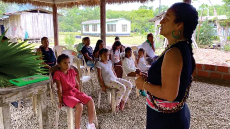 Mujer de pie explicando a niños y adultos sentados bajo un techo (rol de sabedores).