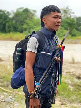 Joven de la guardia con chaleco, morral y bastón de mando en una playa de río (Jóvenes).