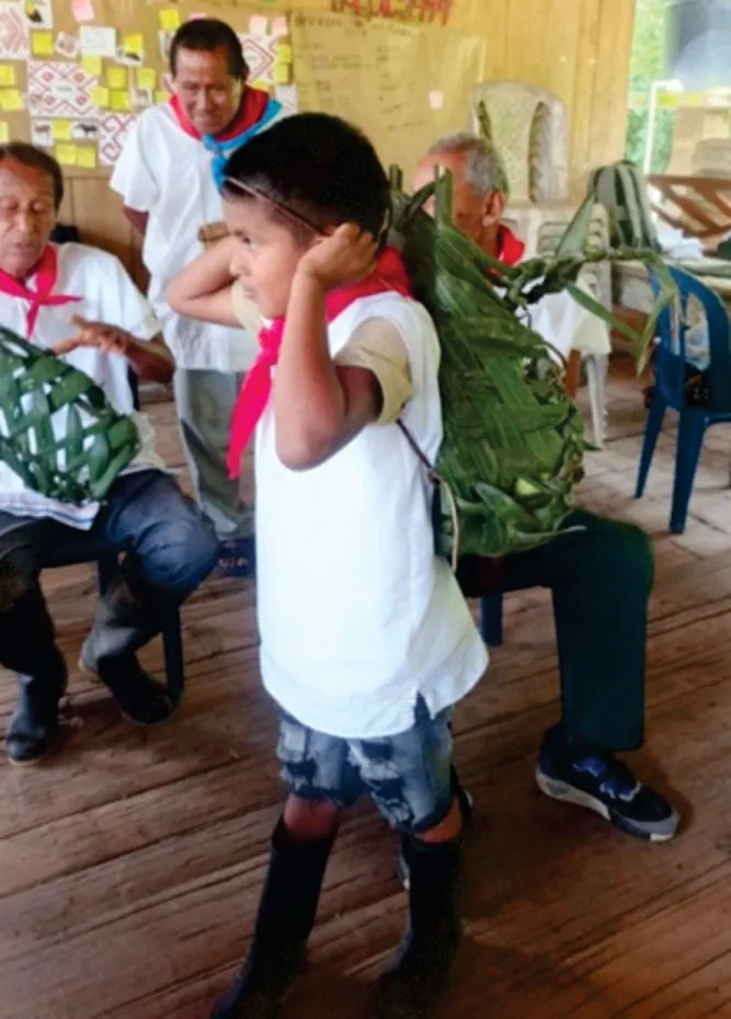 Niño con camisa blanca y pañuelo rojo cargando un canasto de hojas, con mayores sentados alrededor (Formación de liderazgos juveniles).