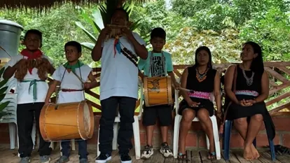 Niños con bombo, flauta y collares, y dos mujeres sentadas, en el salón (Propósito del Plan de Acción).