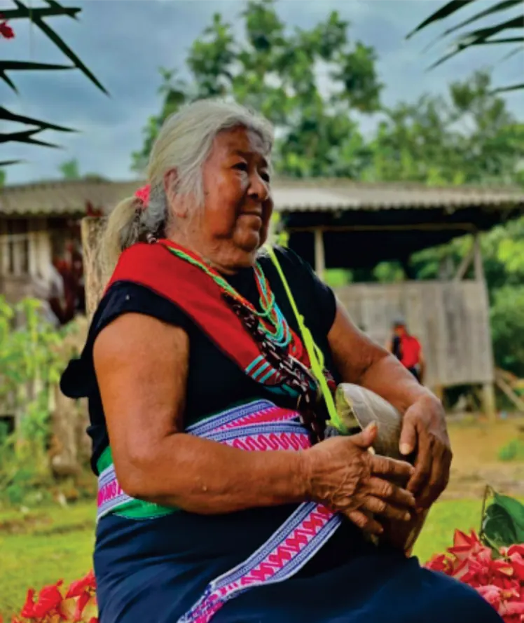Mayora sentada en el patio, con blusa de colores, collares de chaquira y faja tejida, frente a una casa de madera.