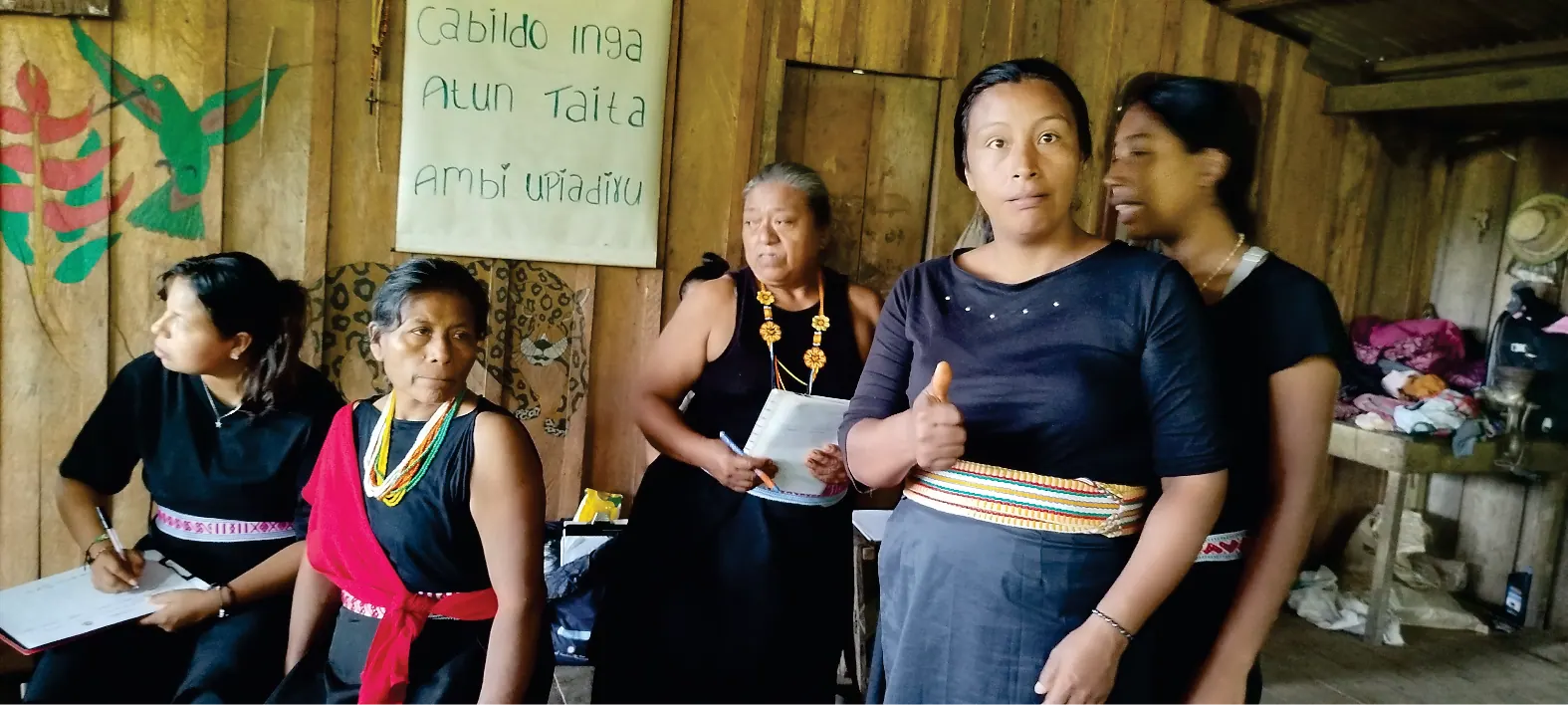 Mujeres con traje propio de pie en el interior de una casa de madera, en un espacio de reunión.