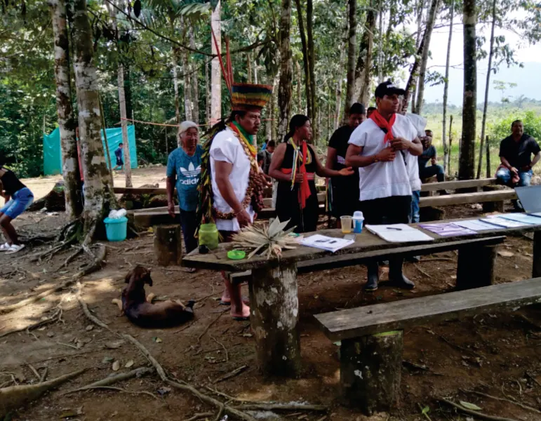 Espacio abierto con mesas de madera donde se reúnen personas, entre ellas un taita con corona de plumas.