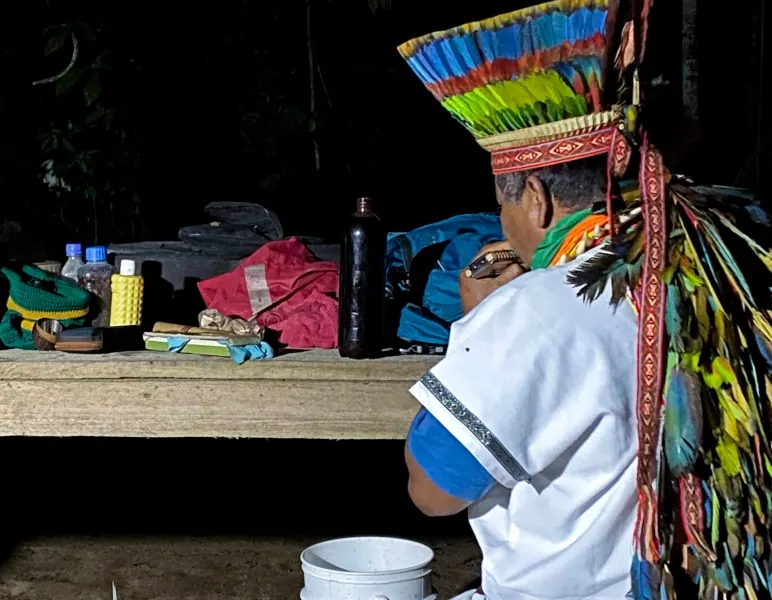 Taita con corona de plumas sentado ante una mesa con elementos de ceremonia, en un espacio oscuro (Ambi Wasi).