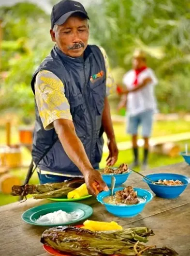Hombre con gorra que sirve alimentos tradicionales sobre una mesa con hojas y recipientes.