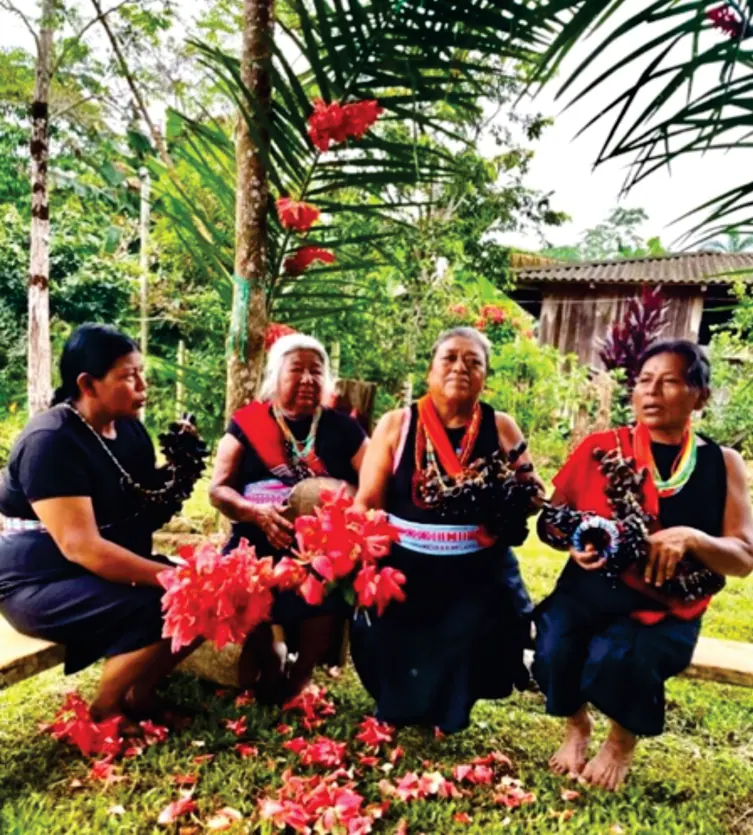 Mujeres sabedoras con traje propio sentadas en el patio con flores rojas.