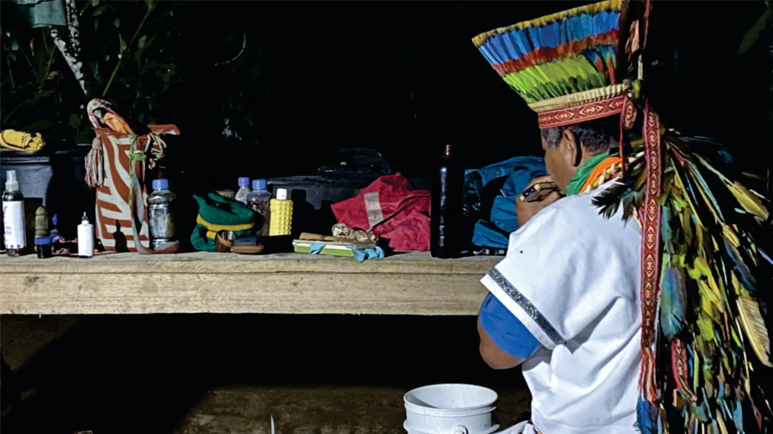 Taita con corona de plumas preparando una ceremonia de noche ante una mesa con elementos rituales.