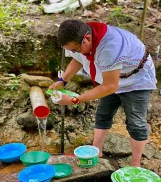 Hombre con pañoleta roja que recoge agua de una manguera en un nacimiento, con recipientes de colores.