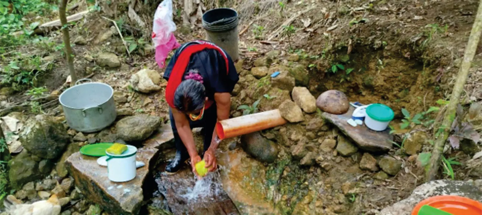 Mujer que lava en una fuente de agua entre piedras, con recipientes.
