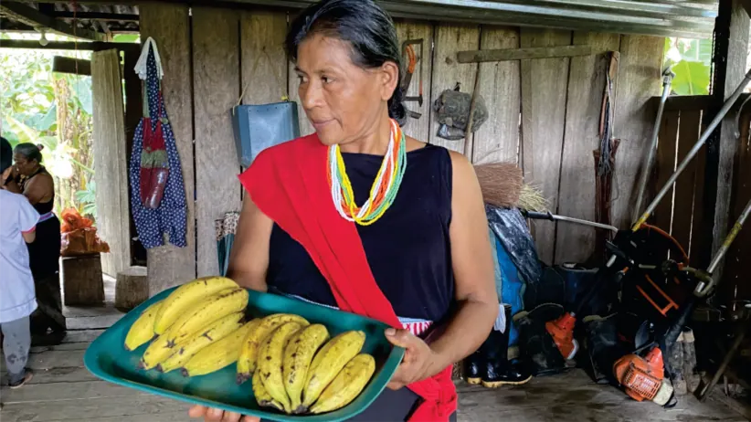 Mujer con traje propio y collares que sostiene una bandeja de plátanos (registro fotográfico).