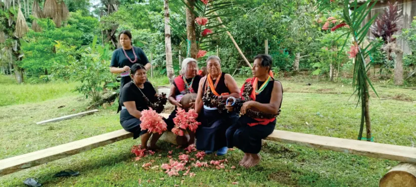 Mujeres sentadas en un tronco con flores rojas, en el patio (registro fotográfico).