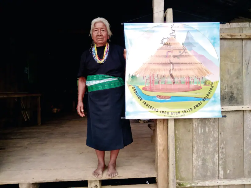 Mayora con traje propio de pie junto a un cartel con el escudo del cabildo (registro fotográfico).