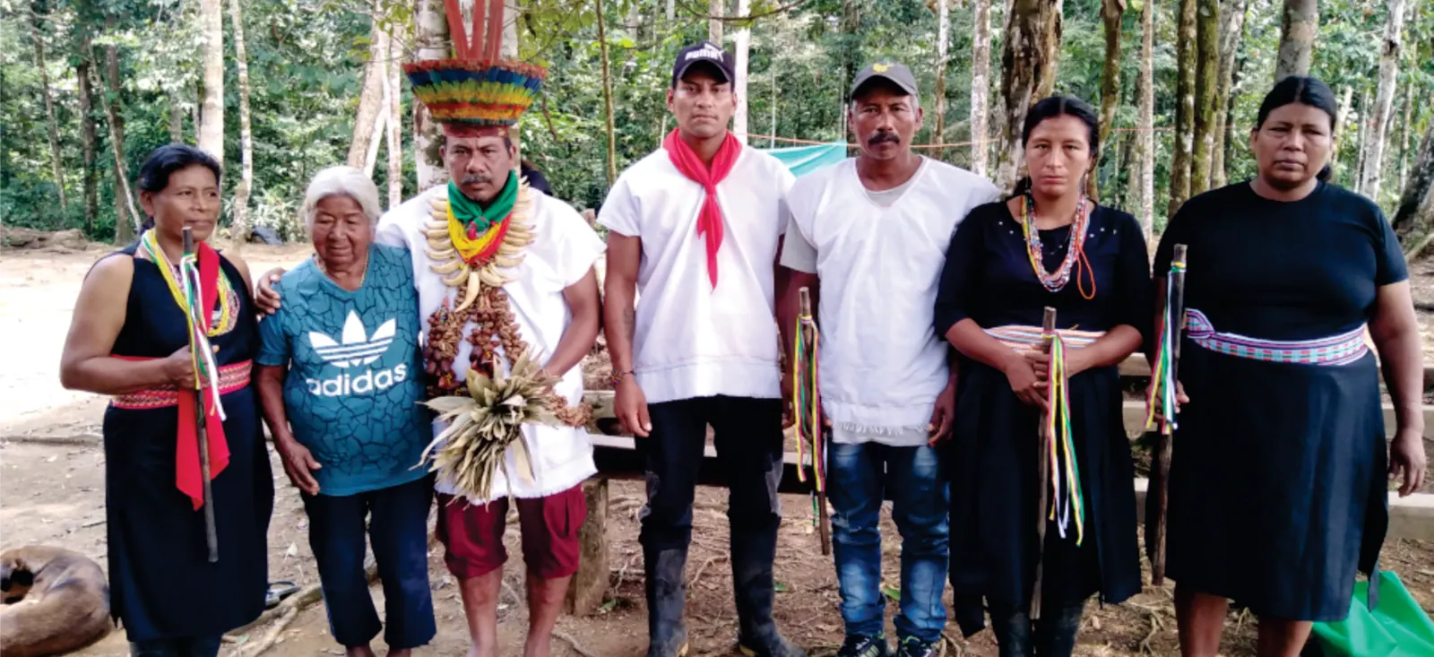 Siete personas de la comunidad en el bosque, una con corona de plumas (registro fotográfico; variante de la foto de p5).