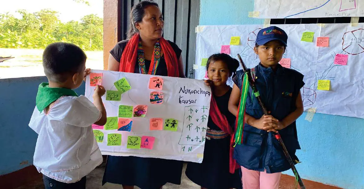 Niños con chaleco de la guardia y bastón, y una mujer, presentando una cartelera «Nukanchipa Kausai» con notas adhesivas.