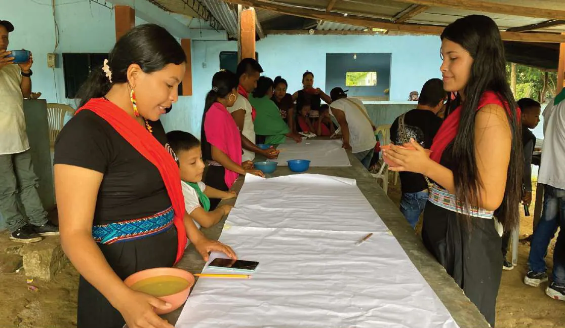 Mujeres con traje propio y pañoleta roja que sirven alimentos junto a un papelógrafo extendido sobre una mesa larga.