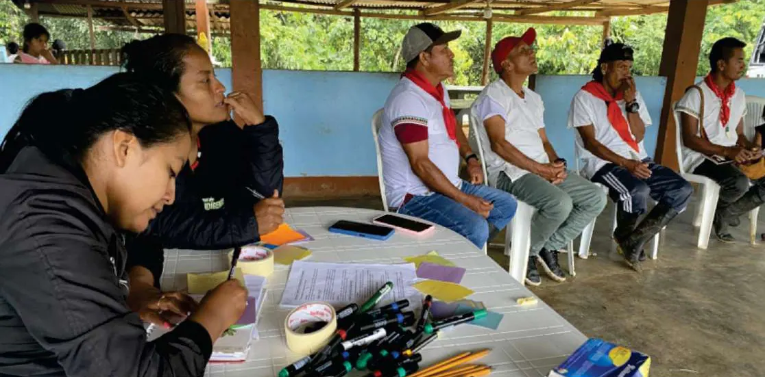 Mujer que escribe en una mesa con marcadores, junto a hombres con pañoleta roja sentados.