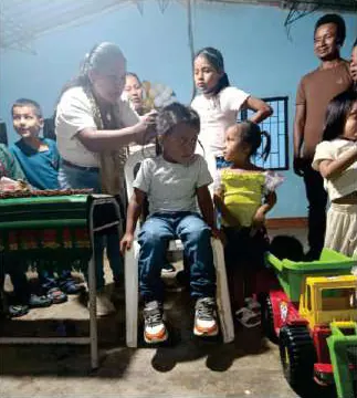 Mayora que corta el pelo a una niña sentada, rodeada de niños y adultos (corte de pelo).