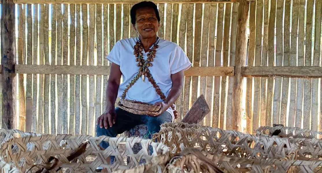 Mayor con collares cruzados sentado detrás de tejidos de bejuco (artesanías).
