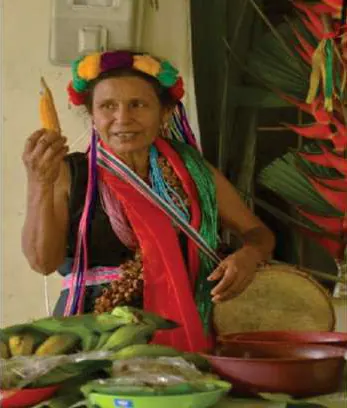 Mayora con corona de flores y collares, con chal rojo, sosteniendo una mazorca.