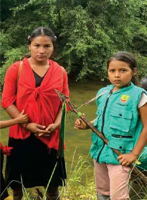 Mujer con chal rojo y niña con chaleco de la guardia y lanza, junto a un río.
