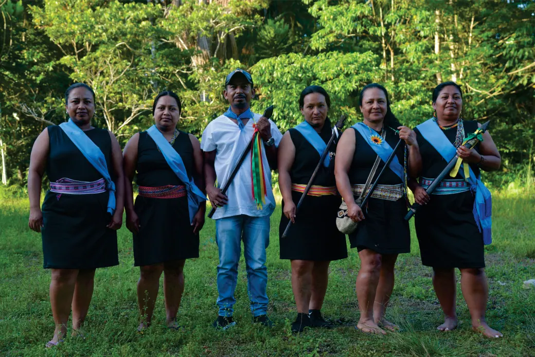 Seis personas de la comunidad (cinco mujeres con banda azul y falda negra, y un hombre) de pie en un potrero junto al bosque.
