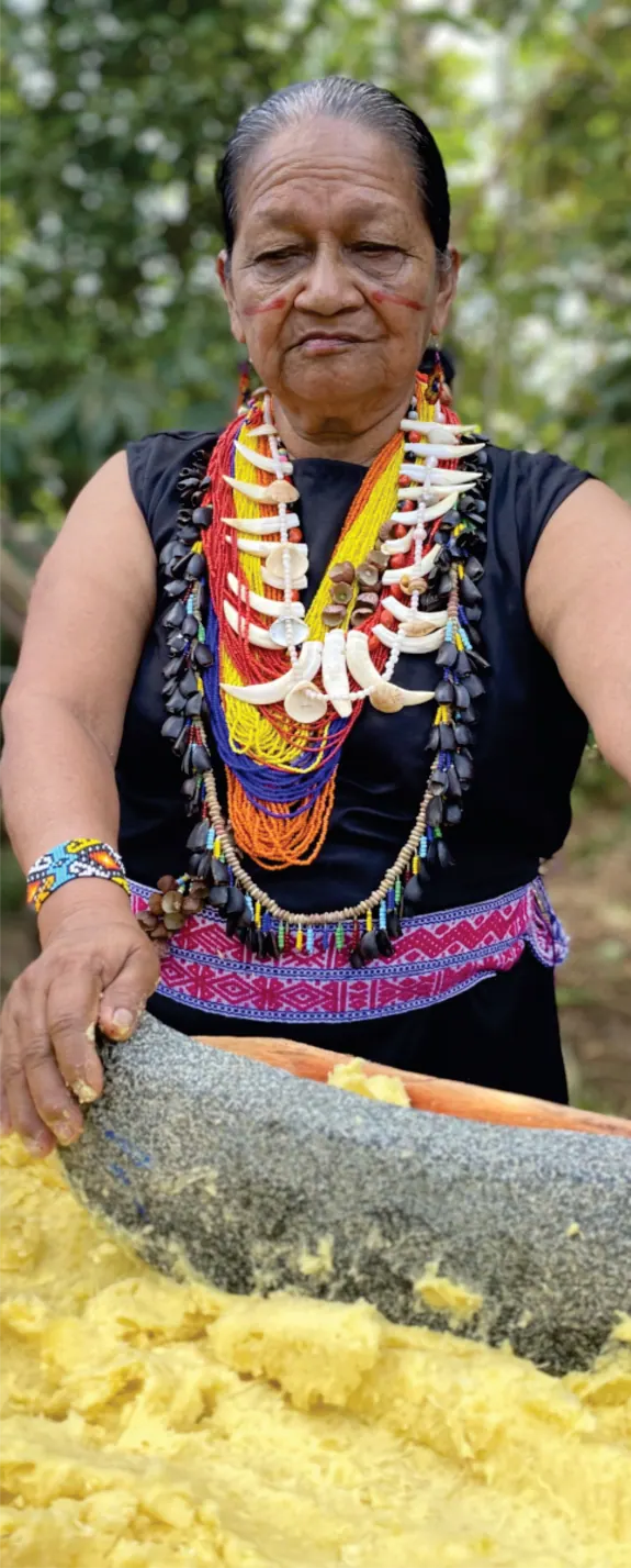 Mayora con collares de chaquira y colmillos que muele sobre una piedra (preparación de alimentos).