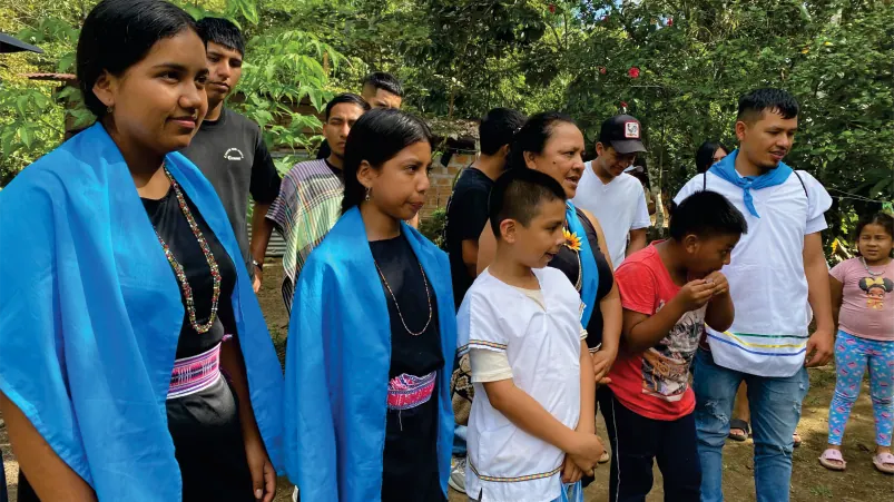 Jóvenes con banda azul y niños de pie junto al bosque.