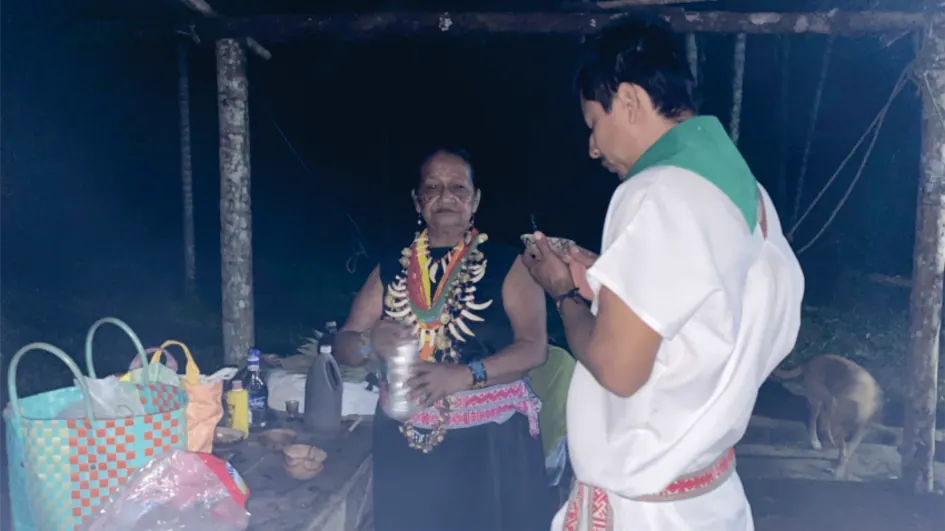 Sabedora con collares que atiende de noche a un joven con pañoleta verde, junto a una mesa con elementos de ceremonia.