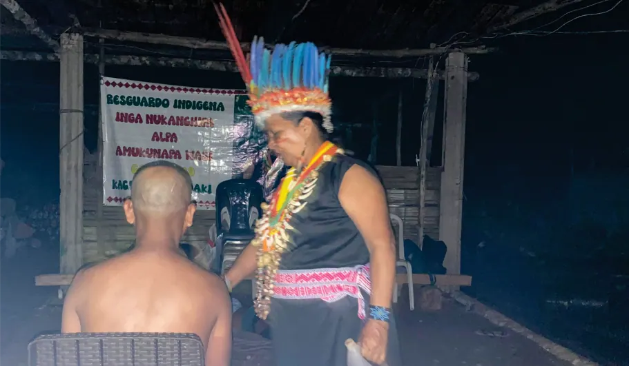 Sabedora con corona de plumas que realiza una curación a un hombre sentado de espaldas, de noche, frente a la pancarta del resguardo.