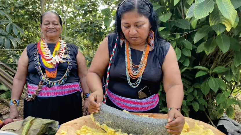 Dos mujeres con collares que muelen sobre una piedra en una batea (preparación de alimentos).