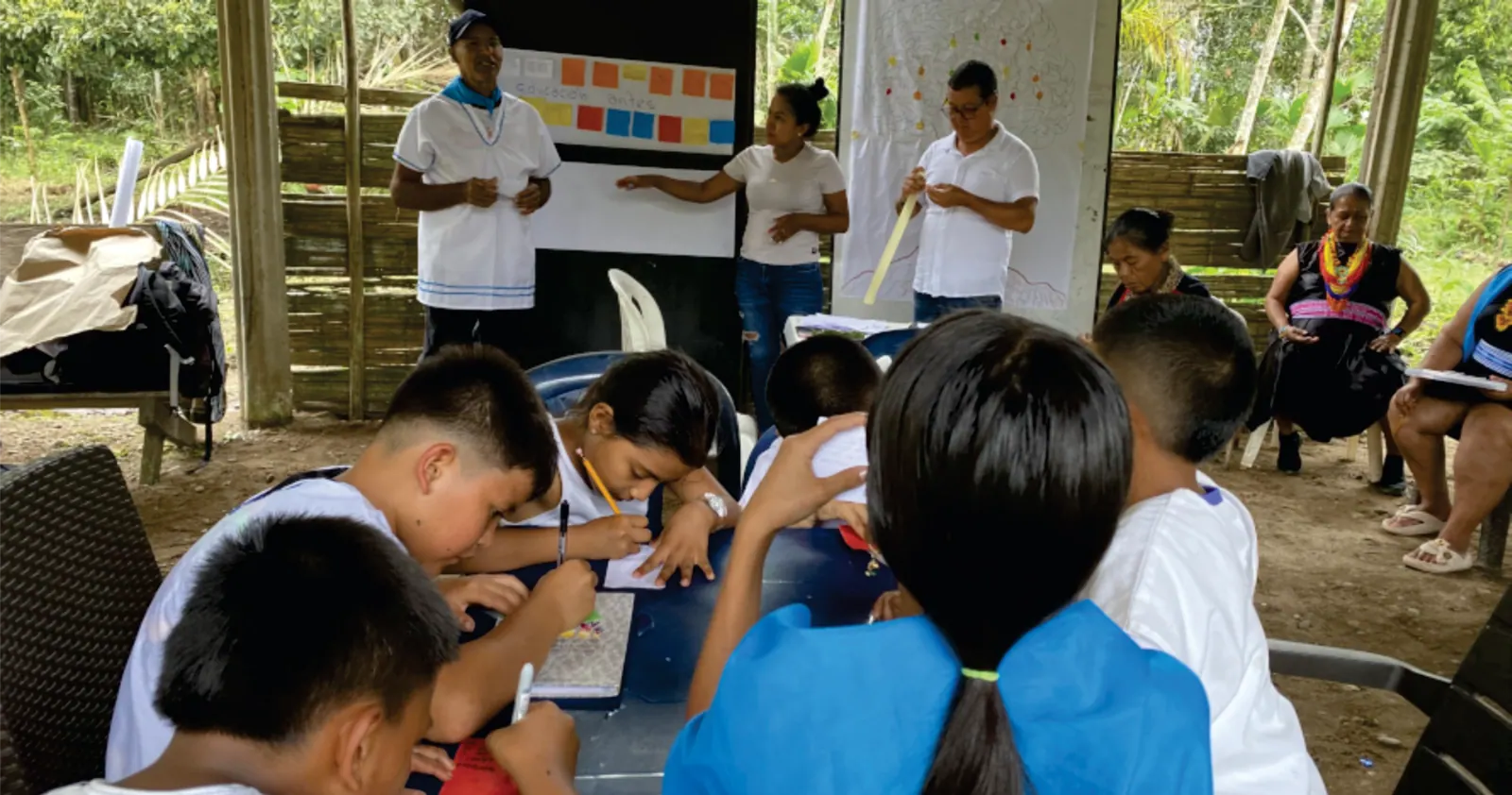 Niños que escriben en mesas durante un taller, con adultos al frente.