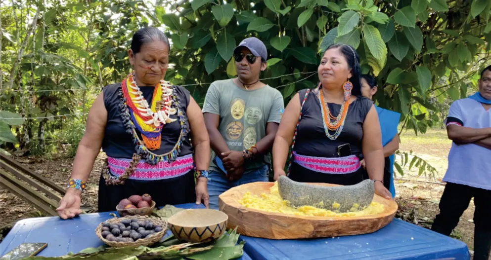 Mujeres con collares que preparan alimentos en una batea, con un hombre sentado al fondo (registro fotográfico).