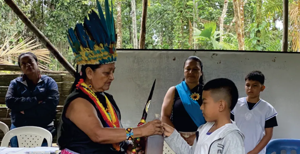 Sabedora con corona de plumas frente a un niño con banda azul durante una actividad (registro fotográfico).