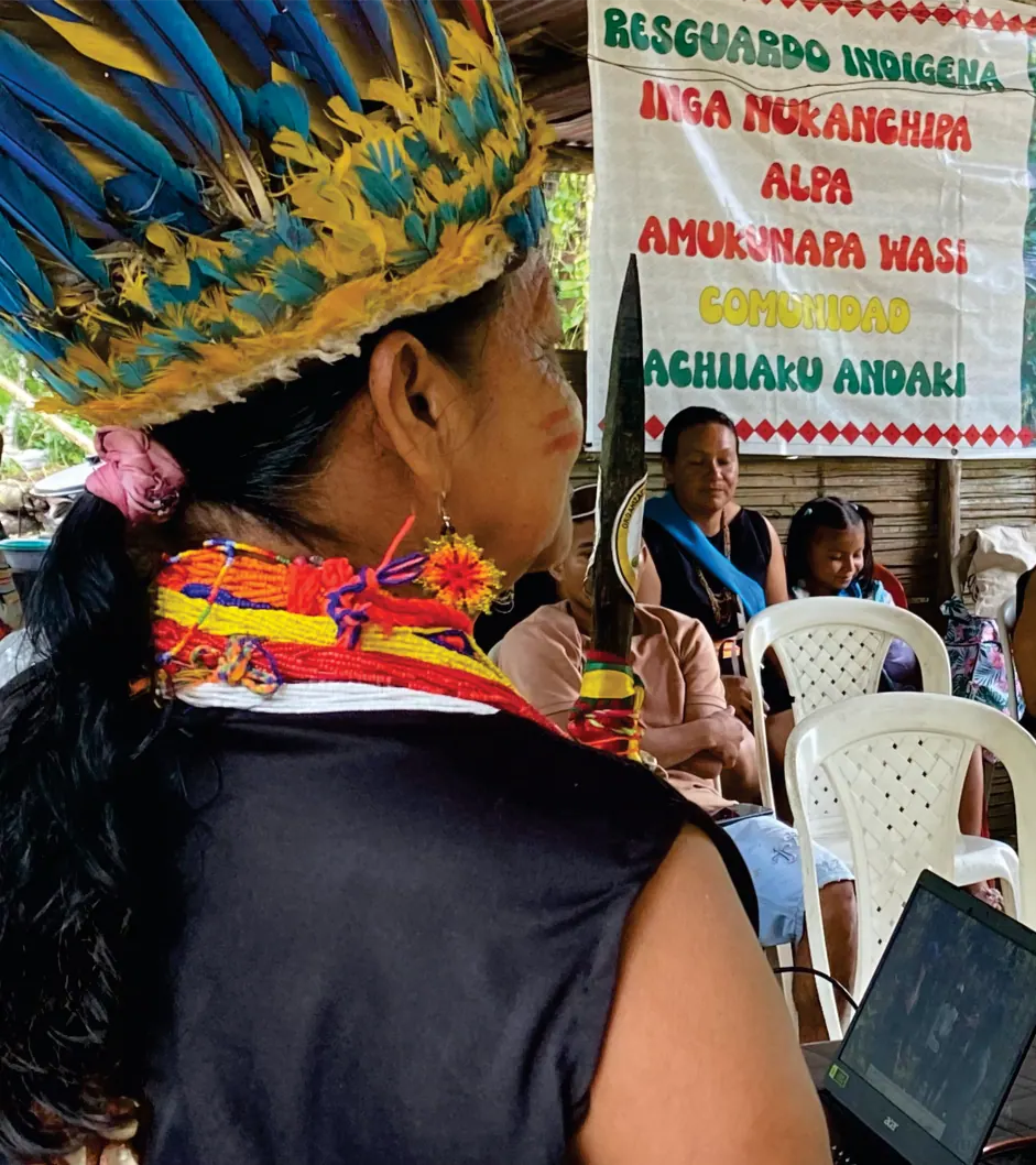Sabedora con corona de plumas, de espaldas, frente a la pancarta del Resguardo Indígena Inga Nukanchipa Alpa Amukunapa Wasi, Kachiiaku Andaki (registro fotográfico).