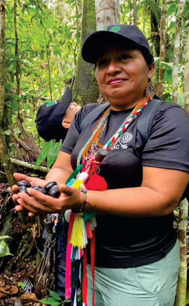 Mujer con gorra y collares que sostiene semillas en el bosque (registro fotográfico).