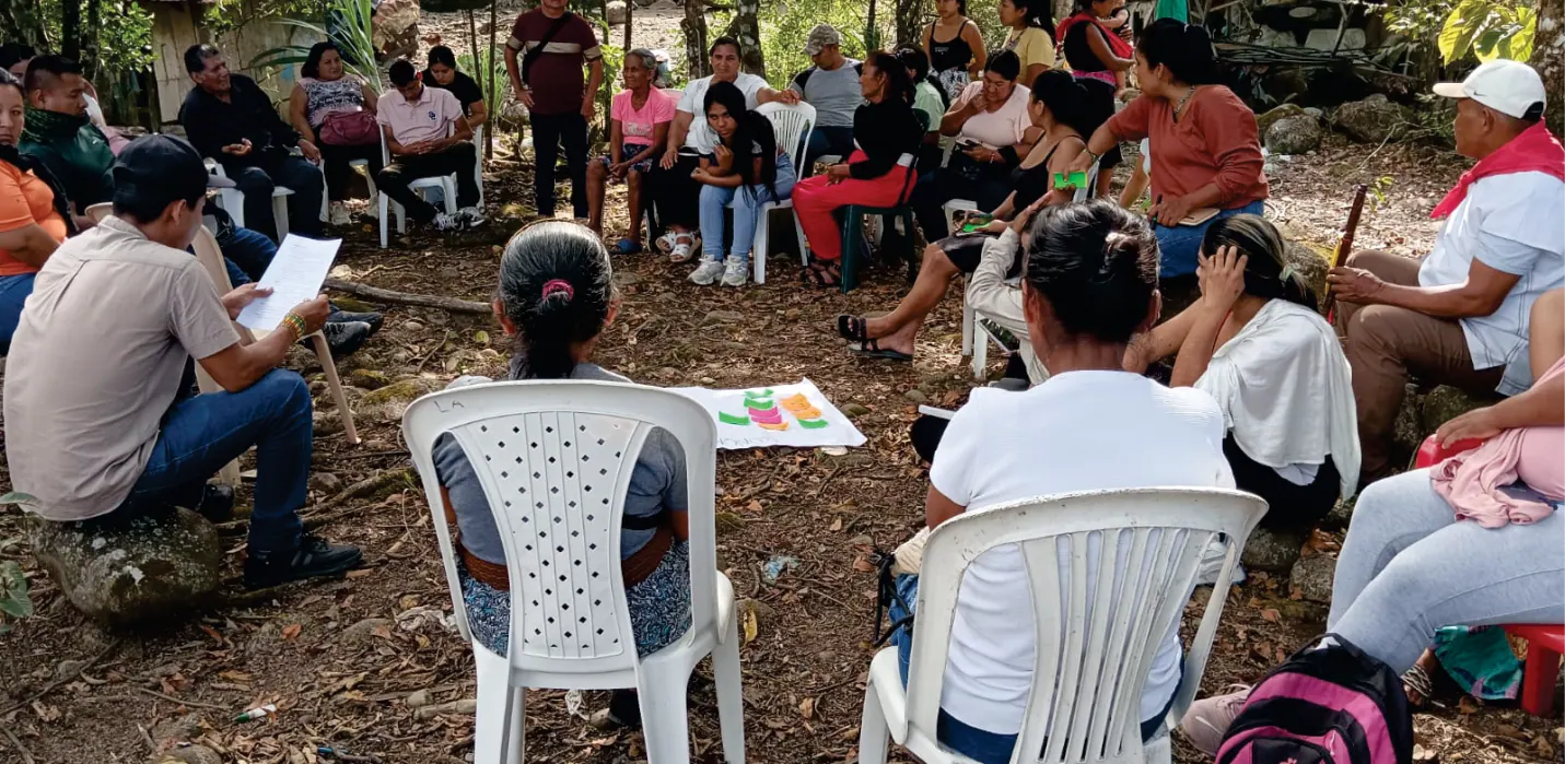 Reunión en círculo al aire libre bajo los árboles, con personas sentadas en sillas plásticas y papeles en la mano.