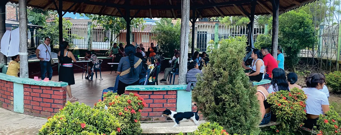 Encuentro comunitario en un kiosco con murete de ladrillo; personas sentadas y plantas ornamentales al frente.