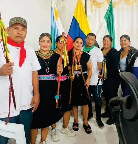 Autoridades con bastones de mando y pañoletas rojas, y mujeres con traje tradicional y collares, frente a banderas.