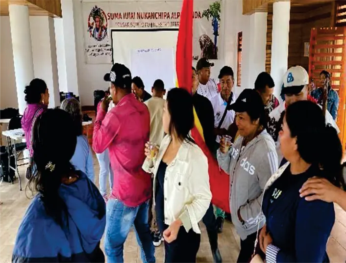 Asamblea en un salón: personas de pie conversando; al fondo un pendón y una bandera.