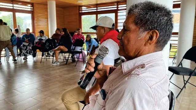Mayor con pañoleta roja hablando en un salón de reuniones; personas sentadas al fondo.